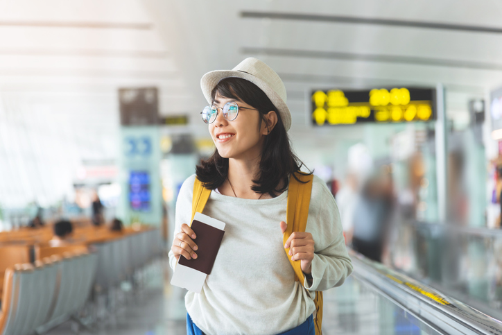 Asian woman wear glasses, hat with yellow backpack is holding flying ticket, passport at the hall of airport.